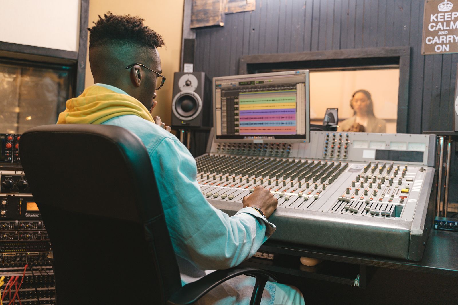 A music producer at a mixing console in a recording studio, a singer visible through the glass